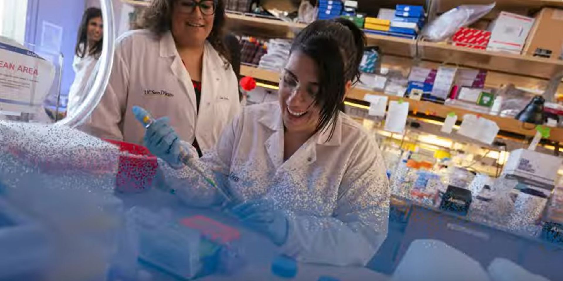 two lab workers watch a third worker using a long pipette