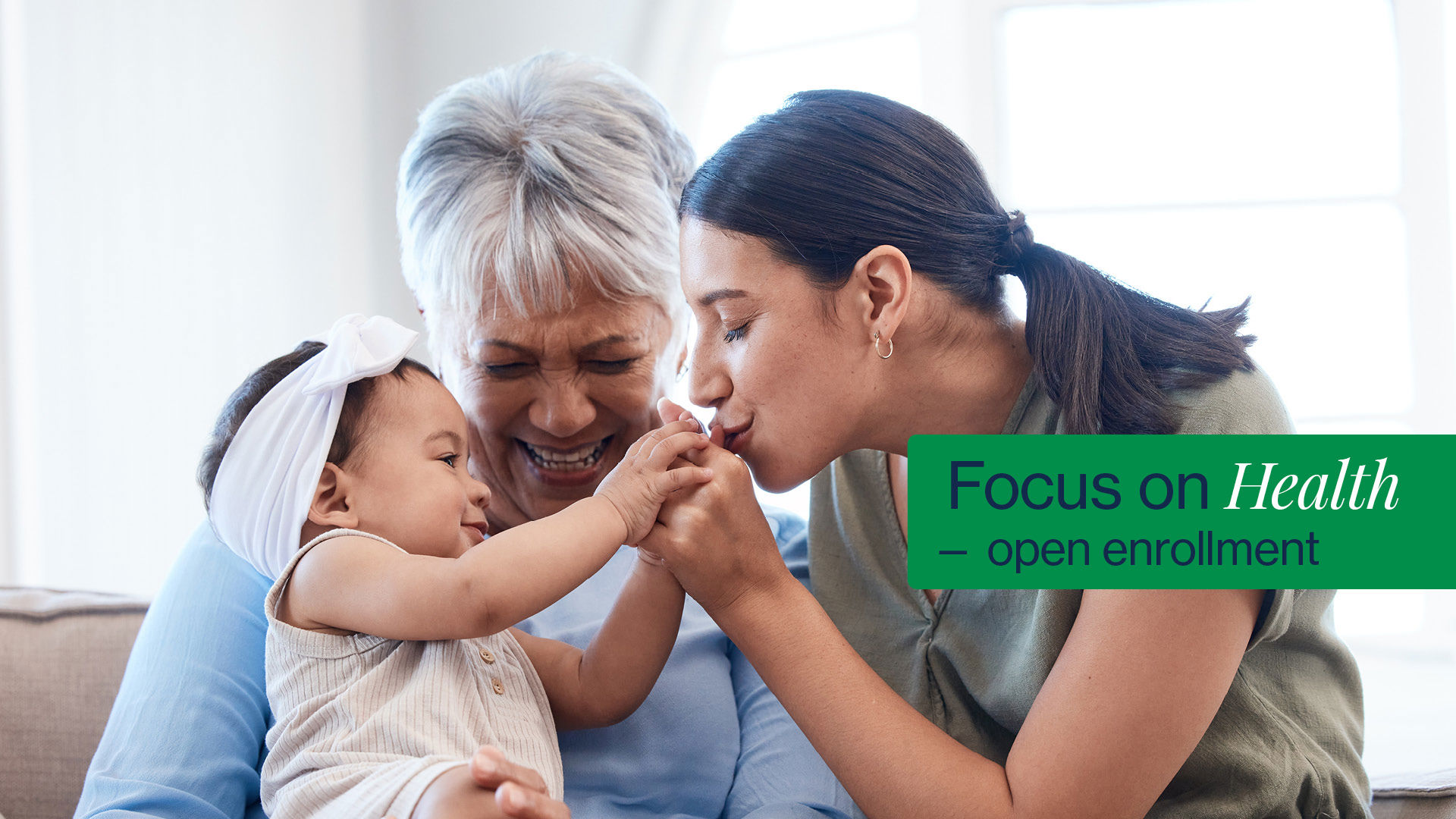 Older woman holds toddler whose hands are being kissed by mom. Shows a graphic with the words "Focus on Health – open enrollment."