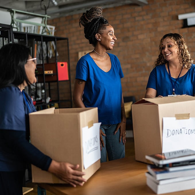 Woman holding box labeled donations laughs and talks with two other volunteers in a store room.