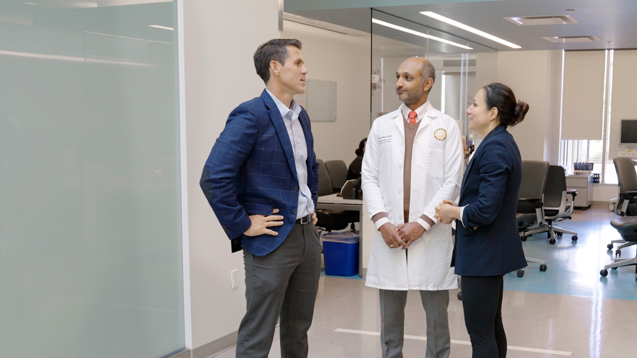 from left, two male doctors have discussion with female doctor standing in front of glass-paned medical office